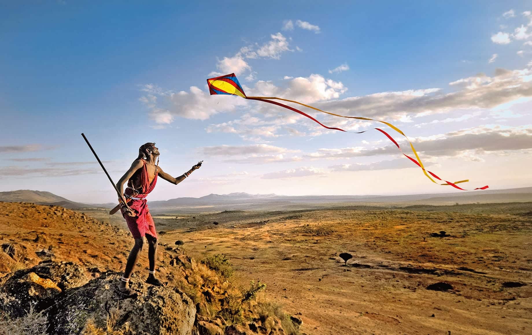 A Maasai warrior flies a kite over a valley with a beautiful landscape in background. Kenya, Africa.